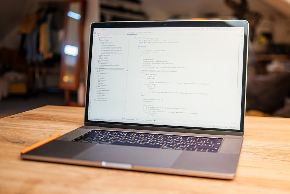 A silver laptop with a black keyboard is placed on a wooden table in a well-lit room, displaying lines of coding on its screen. The background features a cozy, slightly blurred interior with warm ligh