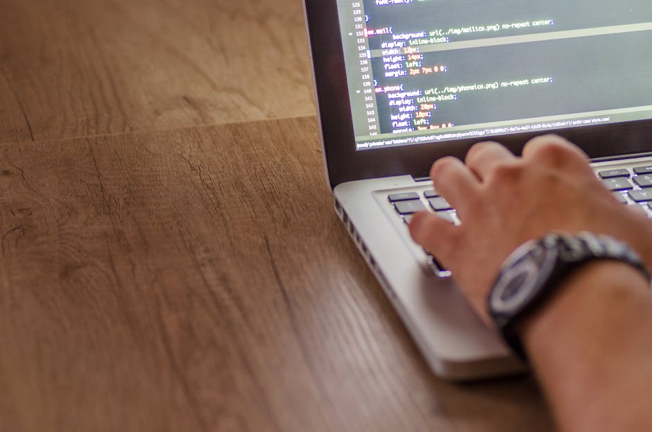 A silver laptop with a black keyboard is placed on a wooden table in a well-lit room, displaying lines of coding on its screen. The background features a cozy, slightly blurred interior with warm ligh