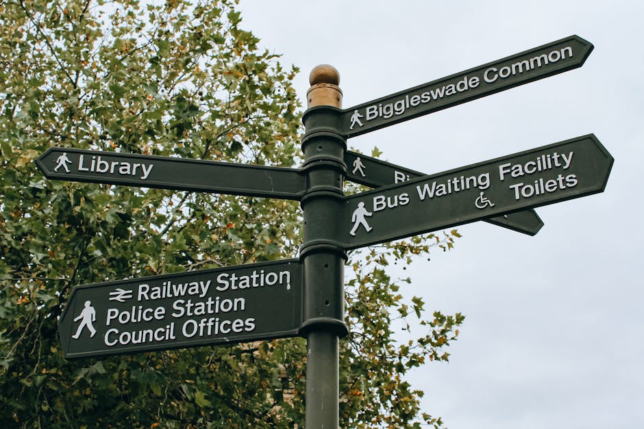 A black metal directional signpost featuring multiple rectangular signs with white text and icons, set against a backdrop of leafy trees and an overcast sky. The signs point to various local amenities